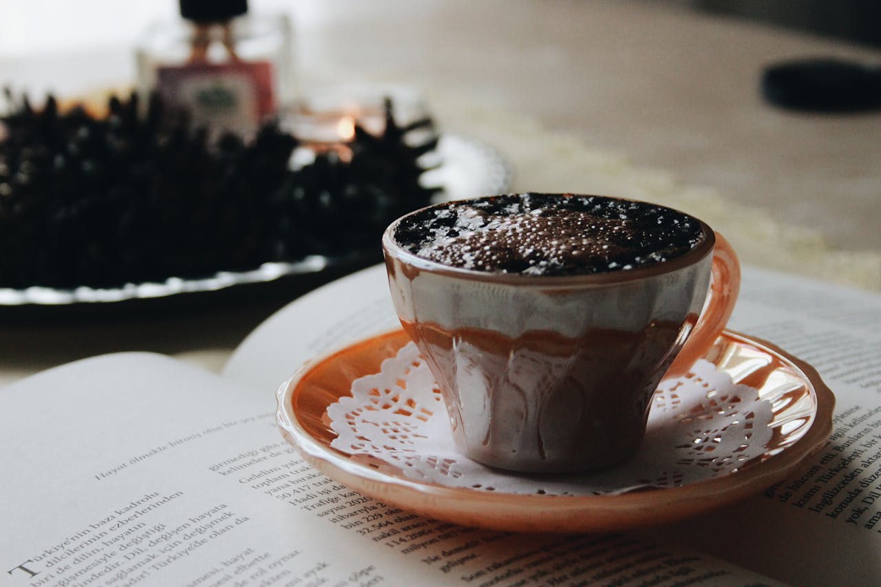 Offerings Cozy scene of a cappuccino on a saucer, open book, and pinecones, perfect for autumn vibes.